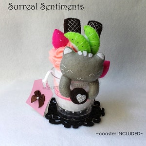 May include: A grey felt cat with pink cheeks and a heart-shaped cookie is sitting in a clear glass jar with a pink and green felt topping. The jar is on a black lace coaster. The text "Surreal Sentiments" is at the top of the image and "coaster INCLUDED" is at the bottom.