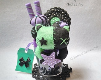 May include: A green and black felt cat with a star and an X on its face, surrounded by purple and green felt candy, a black and white cookie, and a purple and white star. The cat is in a clear glass with a black lace coaster. The text "Surreal Sentiments by Andrea Ng" is at the top of the image. The text "coaster included" is at the bottom of the image.