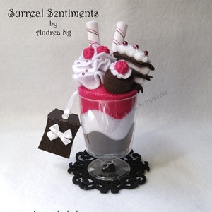 Pode incluir: Um sundae de sobremesa de feltro com uma base em camadas rosa, branca e marrom, coberto com uma colherada de chantilly branco, uma bola de chocolate e dois palitos de wafer cobertos de chocolate. O sundae está em um copo transparente com um descanso de copo preto.