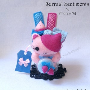 May include: A pink and blue felt cat plush toy in a clear glass jar with a blue and white felt cupcake, pink and white felt waffle, and a blue and white felt macaron. The jar is sitting on a black coaster. The text "Surreal Sentiments by Andrea Ng" is at the top of the image. The text "© Surreal Sentiments" is on the right side of the image. The text "~coaster included~" is at the bottom of the image.