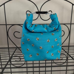 May include: A bright turquoise lunch bag featuring a repeating pattern of small yellow and black bees. The bag has a roll-top closure secured with a black plastic side-release buckle, creating a handle for carrying the rectangular fabric container.