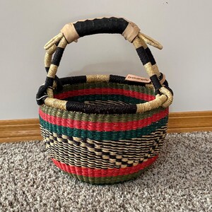 May include: A round woven basket featuring horizontal bands of red, green, and black, with a natural tan rim. The basket has a single arched handle wrapped in black material with tan accents and a small rectangular leather tag attached to the rim.