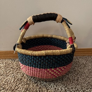 May include: A round woven basket featuring a vibrant colour palette of teal, hot pink, and natural straw tones. The basket includes a sturdy handle wrapped in dark leather at the centre, with additional red and teal woven accents near the base.