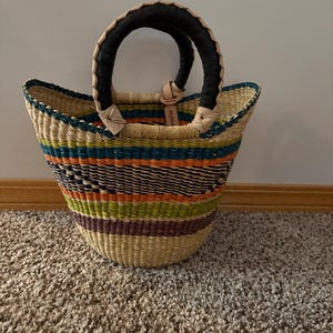 May include: A woven basket bag featuring horizontal stripes in shades of orange, green, blue, and dark purple. The bag has two rounded handles wrapped in black material with light tan leather accents and a small leather tag attached to one handle.