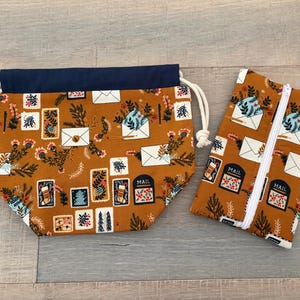 May include: A fabric drawstring pouch with a navy blue top and a mustard yellow base featuring a holiday mail print, alongside a matching rectangular zippered pouch. Both items display illustrations of envelopes, mailboxes, and pine branches.