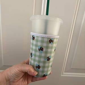 May include: A clear plastic tumbler with a lid and a dark green straw, featuring a removable fabric sleeve. The sleeve has a light green and white gingham check pattern decorated with small yellow and black bees with blue wings.