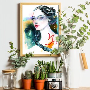 May include: Watercolour painting of a woman with a Capricorn zodiac symbol on her chest. The painting is framed in a light wood frame and is surrounded by plants and a camera.