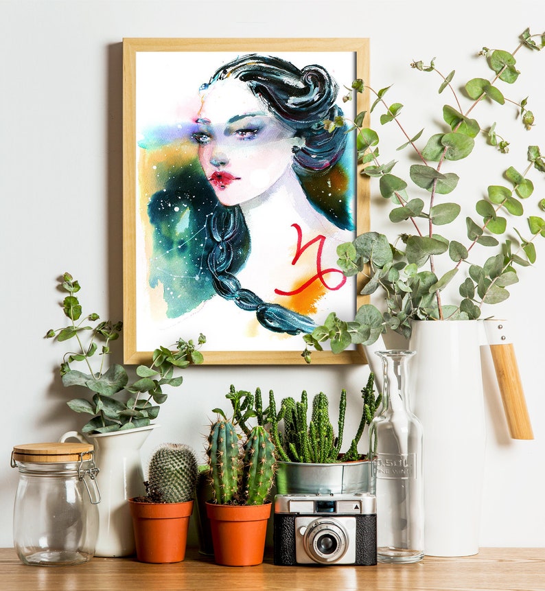 May include: Watercolour painting of a woman with a Capricorn zodiac symbol on her chest. The painting is framed in a light wood frame and is surrounded by plants and a camera.