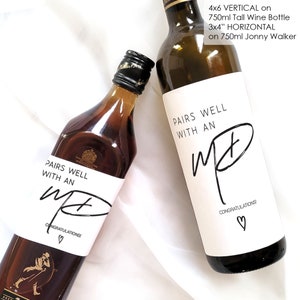May include: Two wine bottle labels with a white background and black text. The text on both labels reads "Pairs well with an M&" with a heart symbol at the bottom. The label on the left is on a brown bottle and the label on the right is on a clear bottle. The label on the right also includes the text "Congratulations!"