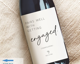 May include: A white wine bottle label with black text that reads "Pairs well with getting engaged."  The label also includes the text "Congratulations!" and "Kelly & Tom" with a small heart symbol.