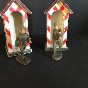 May include: Two toy soldiers standing in wooden guard booths. The booths are painted white with red stripes. The soldiers are wearing green uniforms and holding rifles.