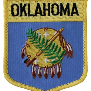 May include: Embroidered patch featuring the word "OKLAHOMA" in black block letters above a shield design. The shield has a blue background with a central emblem of a yellow Native American shield, a peace pipe, and olive branches. The patch is bordered in yellow.