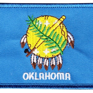 May include: A blue embroidered patch with the state seal of Oklahoma. The seal features a yellow circle with a green sprig of  Indian grass, a crossed tomahawk and peace pipe, and seven white feathers. The word "OKLAHOMA" is embroidered in white below the seal.