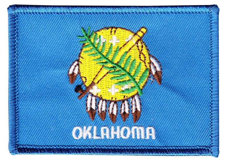 May include: A blue embroidered patch featuring the state seal of Oklahoma. The seal depicts a yellow circle with a green sprig of Indian grass, a crossed tomahawk and peace pipe, and seven white feathers. The word "OKLAHOMA" is embroidered in white below the seal.