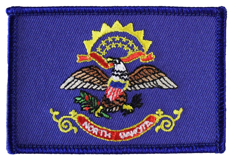 以下が含まれることがあります： ノースダコタ州の州旗の刺繍パッチ。旗は青色で、13個の星がついた金色の太陽の光、赤いリボン、赤、白、青の盾を持った金色のワシ、緑色の小麦の穂が描かれています。「NORTH DAKOTA」という文字は赤と金で書かれています。