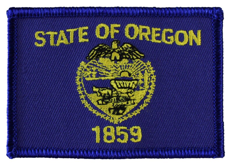 May include: A blue embroidered patch with the words "STATE OF OREGON" and the state seal in yellow. The seal features a beaver, a wagon wheel, and a mountain range. The year "1859" is also embroidered in yellow.