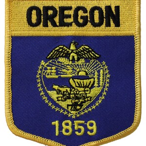 May include: A yellow and blue embroidered patch with the word "OREGON" in black lettering at the top and the state seal in the center. The seal features a golden eagle with a shield containing a wagon, a sunrise, and a mountain range. The year "1859" is printed in yellow below the seal.