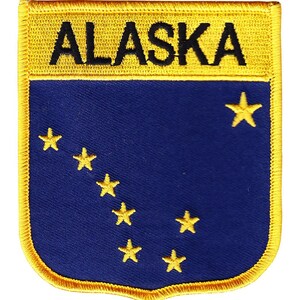 May include: Embroidered patch featuring the word "ALASKA" in black on a gold background. The patch has a blue field with eight gold stars, representing the state flag. The patch has a gold border.