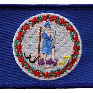May include: A blue embroidered patch featuring the state seal of Virginia. The seal depicts a woman standing on a purple rock, holding a spear and a rifle, with a man lying at her feet. The seal is surrounded by a wreath of red and green leaves.