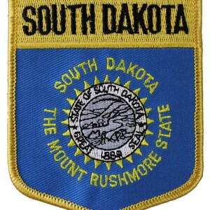 May include: A blue and gold embroidered patch featuring the state seal of South Dakota. The seal depicts a sun with rays, a mountain range, a river, and the words "The Great Seal of the State of South Dakota" and "1889". The patch also includes the words "South Dakota" and "The Mount Rushmore State".