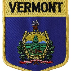 May include: A gold embroidered patch with the word "VERMONT" in black lettering on a yellow background. The patch features the state seal of Vermont, which includes a green pine tree, a blue mountain range, and a red ribbon with the words "VERMONT" and "FREEDOM AND UNITY" in black lettering.
