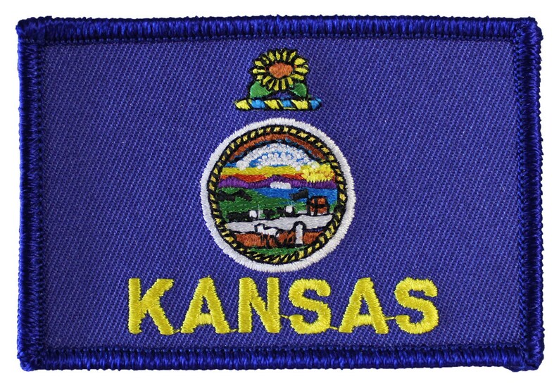 May include: A blue embroidered patch with the state seal of Kansas, featuring a sunflower, a sun, and a river. The word "KANSAS" is written in yellow below the seal.