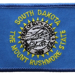 May include: A blue embroidered patch with the state seal of South Dakota. The seal features a sunburst design with the words "The Mount Rushmore State" and "Great Seal of South Dakota" around the edge.