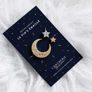 Peut inclure: Un ensemble de trois épingles en émail représentant un croissant de lune et deux étoiles. L'épingle de la lune est dorée avec un motif en émail blanc. Les étoiles sont argentées et roses. Les épingles sont sur une carte bleue avec le texte "Edition Spéciale Le Pins Émaillé L'Encreerie Paris www.lencreerieparis.com".