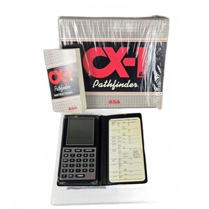 May include: A CX-1 Pathfinder flight computer set. The set includes a black and silver calculator, a booklet, and a larger box, all with the "CX-1 Pathfinder" logo in red. The calculator is open, revealing a screen and buttons. The set is packaged in clear plastic.