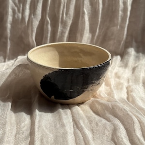 May include: A small, off-white ceramic bowl with a black, curved design on the side. The bowl has a smooth interior and a slightly textured exterior. The bowl is set against a neutral, textured background.