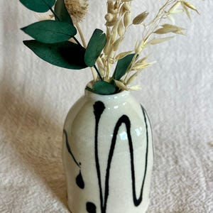 May include: A small, off-white ceramic vase with black dripped paint design, holding a collection of dried flowers and greenery. The arrangement includes green eucalyptus leaves and beige dried grasses, creating a natural, decorative display.