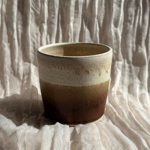 May include: A handmade ceramic cup with a gradient design. The cup transitions from a light beige at the top to a deep brown at the base. The cup is cylindrical and sits on a textured, off-white surface.