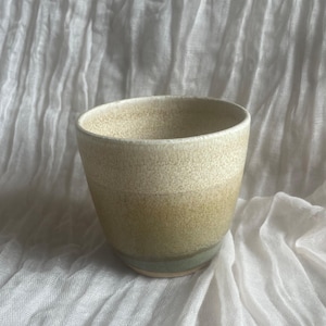 May include: A handcrafted ceramic cup with a rounded form. The cup displays a colour gradient, shifting from a light beige at the top to a muted green at the base. The cup is likely used for drinking beverages.