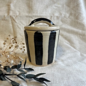May include: A white ceramic jar with a black striped pattern and a black handle on the lid.