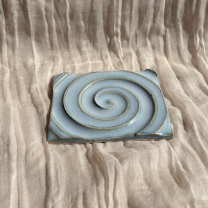 May include: A square, light blue ceramic tile with a spiral design. The spiral is raised and has a gold-colored edge. The tile is a decorative item, possibly for use as a coaster or small art piece.