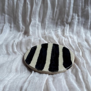 May include: A small, oval ceramic dish featuring a black and white striped design. The dish is set against a white, textured fabric backdrop. The stripes are vertical and evenly spaced, offering a striking contrast.