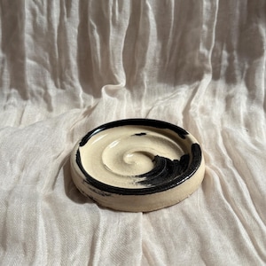 Könnte beinhalten: Eine kleine, runde Keramikschale mit cremefarbenem Innenraum und einem schwarzen Pinselstrich-Design am Rand. Die Schale liegt auf einem weißen, strukturierten Stoff und erzeugt eine minimalistische Ästhetik. Die Schale hat einen Durchmesser von etwa 10 cm.