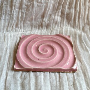 May include: A square, pink ceramic coaster with a spiral design. The coaster is a light shade of pink and has a textured surface. The coaster is approximately 4 inches square.