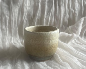 Könnte beinhalten: Eine kleine, handgefertigte Keramiktasse mit abgerundeter Form. Die Tasse weist einen Farbverlauf auf, der von einem hellen Beige oben zu einem dunkleren, erdigen Ton zur Basis übergeht. Die Tasse steht vor einem weißen, strukturierten Stoffhintergrund.