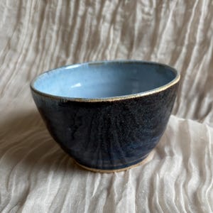 May include: A handmade ceramic bowl with a dark blue exterior and a light blue interior. The bowl has a slightly irregular shape and a textured surface. The rim is a light brown color. This bowl is suitable for serving food or as a decorative item.