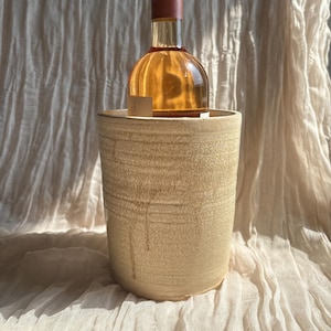 May include: A clear glass wine bottle with a pink label and a black and pink cap is in a beige ceramic wine chiller. The chiller has a textured surface and a cylindrical shape. The background is a light-coloured fabric.