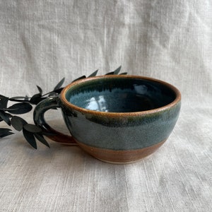 May include: A dark blue ceramic mug with a brown rim and a brown band around the bottom. The mug has a speckled glaze and a rounded shape.