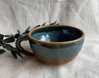 May include: A dark blue ceramic mug with a brown rim and a brown band around the bottom. The mug has a speckled glaze and a rounded shape.