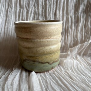 May include: A handmade ceramic cup with a textured, ridged design. The cup features a gradient of colours, starting with a light beige at the top, transitioning to green at the base. The cup is likely used for drinking beverages.