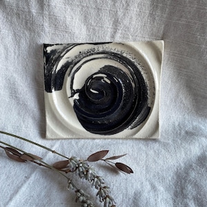 May include: A white ceramic square dish with a black swirl design. The dish is slightly raised around the edges.