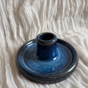 Könnte beinhalten: Ein handgefertigter Kerzenhalter aus Keramik in Blau- und Dunkelgrautönen. Der Kerzenhalter hat einen kreisförmigen Sockel mit einer erhöhten Mitte für die Kerze. Die glänzende Oberfläche verleiht ihm ein modernes Aussehen. Ein dekoratives Wohnaccessoire.