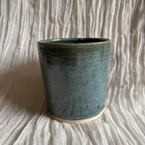 Könnte beinhalten: Ein handgefertigter Keramikbecher mit einer gesprenkelten blau-grünen Glasur. Der zylindrische Becher hat eine leicht ausgestellte Oberseite und einen natürlichen, unglasierten Boden. Der Innenraum ist ein dunklerer Grünton. Die Keramik steht auf einem strukturierten, cremefarbenen Stoff.