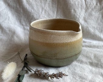 May include: A handmade ceramic bowl with a spout, featuring a gradient of colors from beige to green. The bowl is set on a textured, off-white fabric, with dried lavender and other decorative elements.