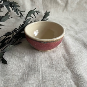 Könnte beinhalten: Eine kleine, cremefarbene Keramikschüssel mit einem roten und braunen Rand. Die Schüssel steht auf einer weißen Stoffoberfläche.
