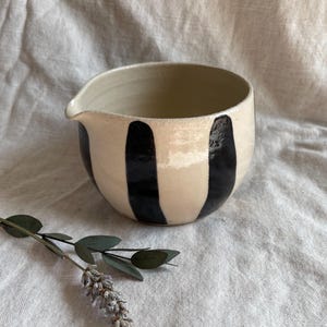 May include: A white ceramic pitcher with black vertical stripes. The pitcher has a rounded shape and a small spout.